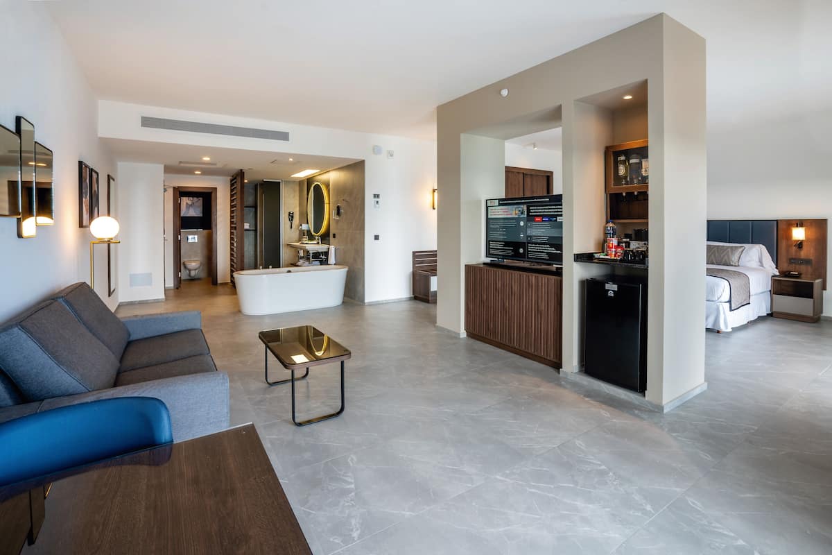 A modern hotel room with a sofa, a coffee table, a kitchen area, and a separate sleeping area.