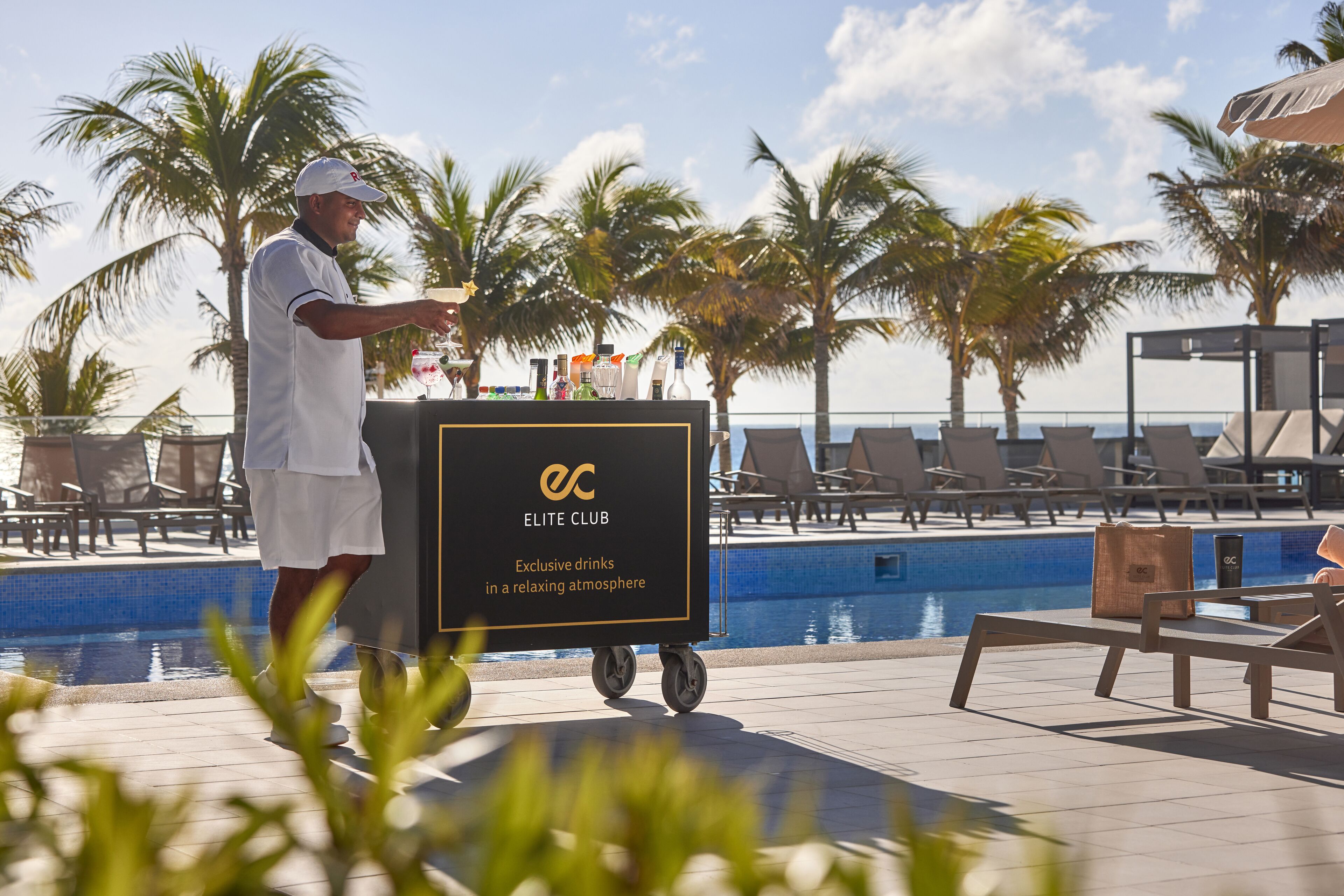 A poolside area with lounge chairs, a man pushing a cart labeled "ELITE CLUB," and palm trees in the background.