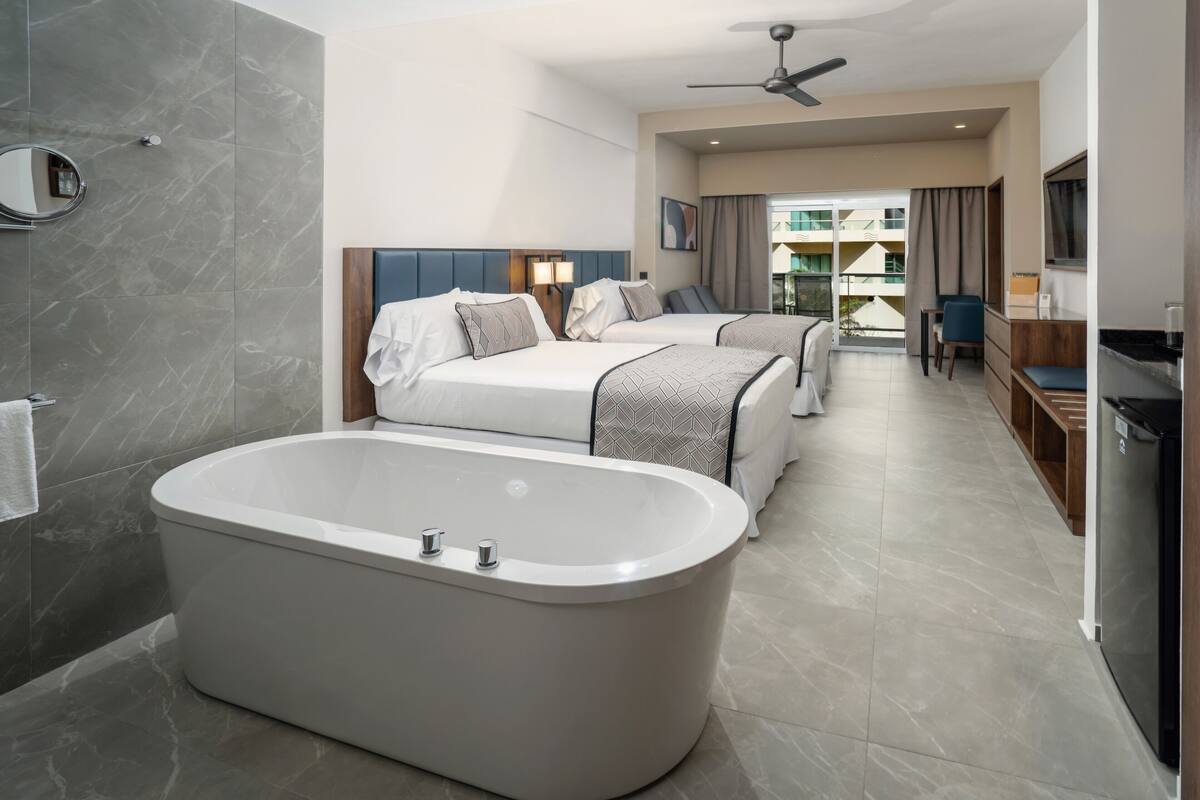 A modern hotel room with a large bathtub, two beds, a desk, and a balcony with a view.