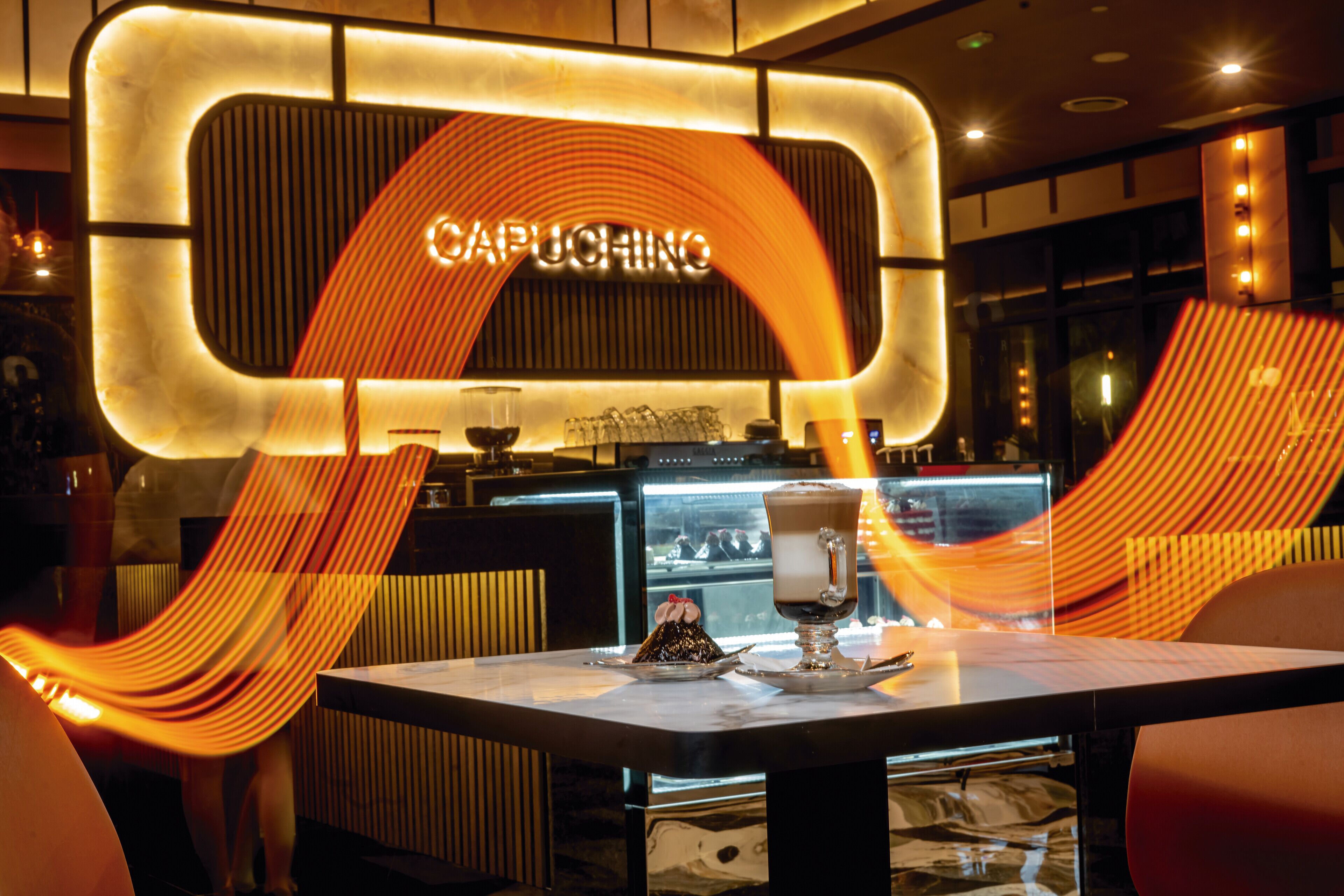 A modern bar area with a prominent 'CAPUCHING' sign, a glass of coffee, and a dessert on a table.