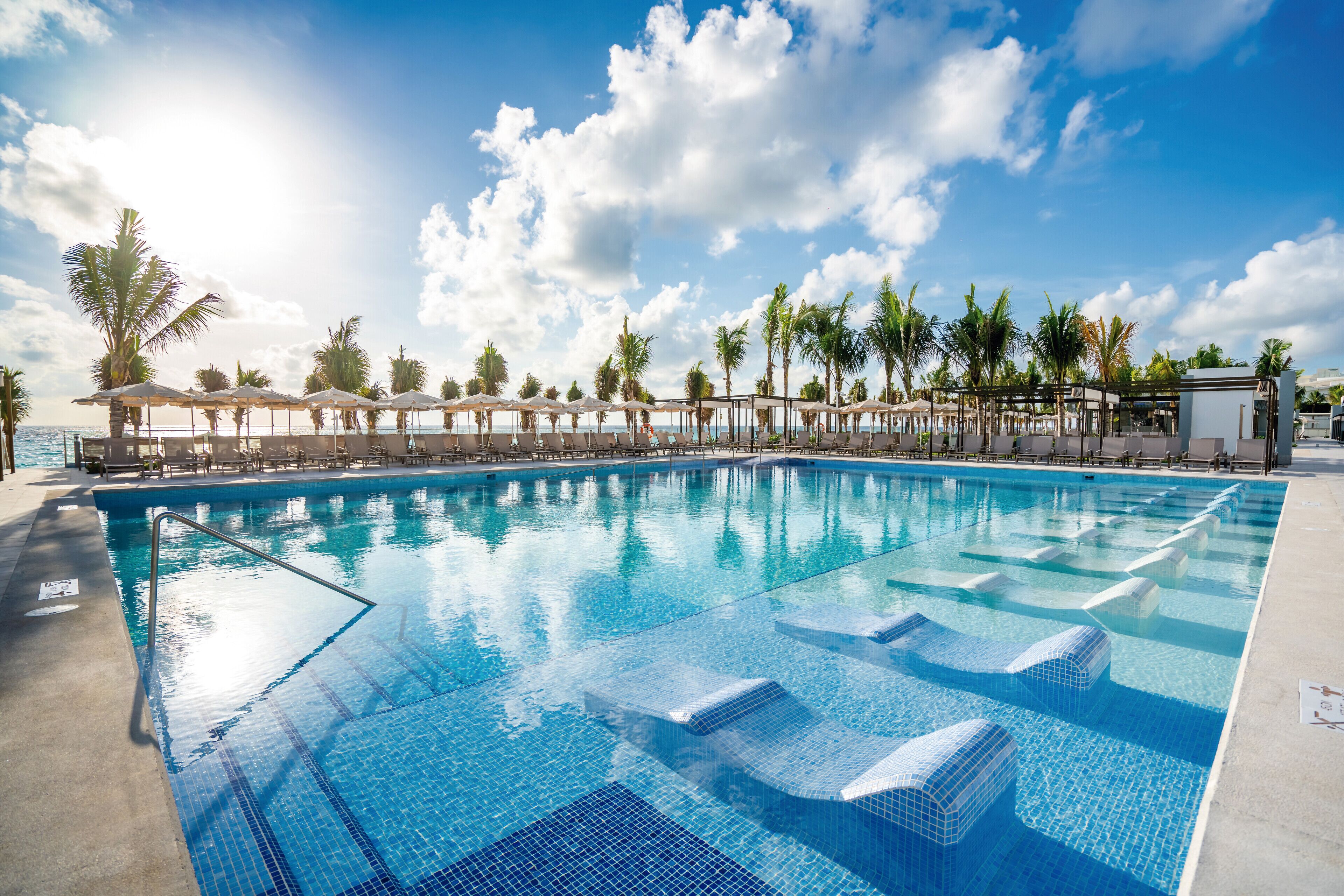 A swimming pool with clear blue water, surrounded by lounge chairs and umbrellas, with palm trees and a clear sky in the background.