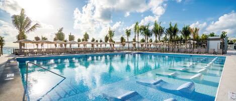 A swimming pool with clear blue water, surrounded by lounge chairs and umbrellas, with palm trees and a clear sky in the background.