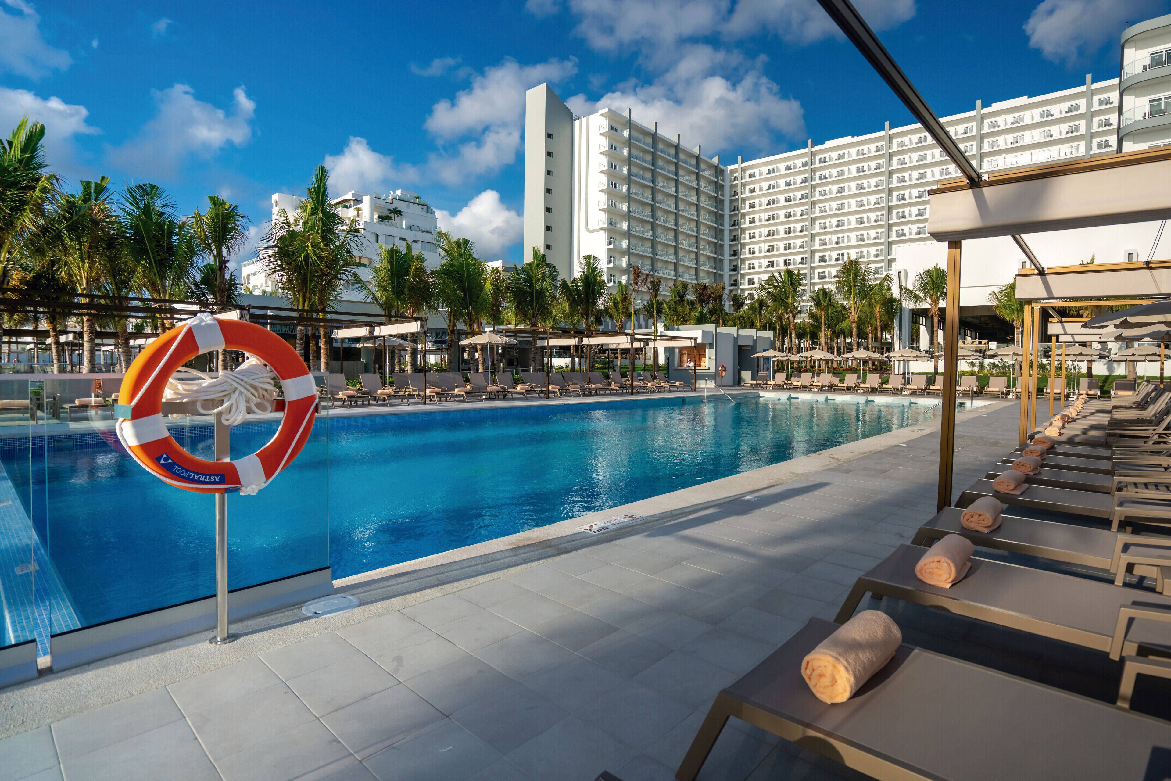 Ein Swimmingpool mit Liegestühlen und einem Rettungsring, umgeben von Palmen und einem Hotelgebäude.