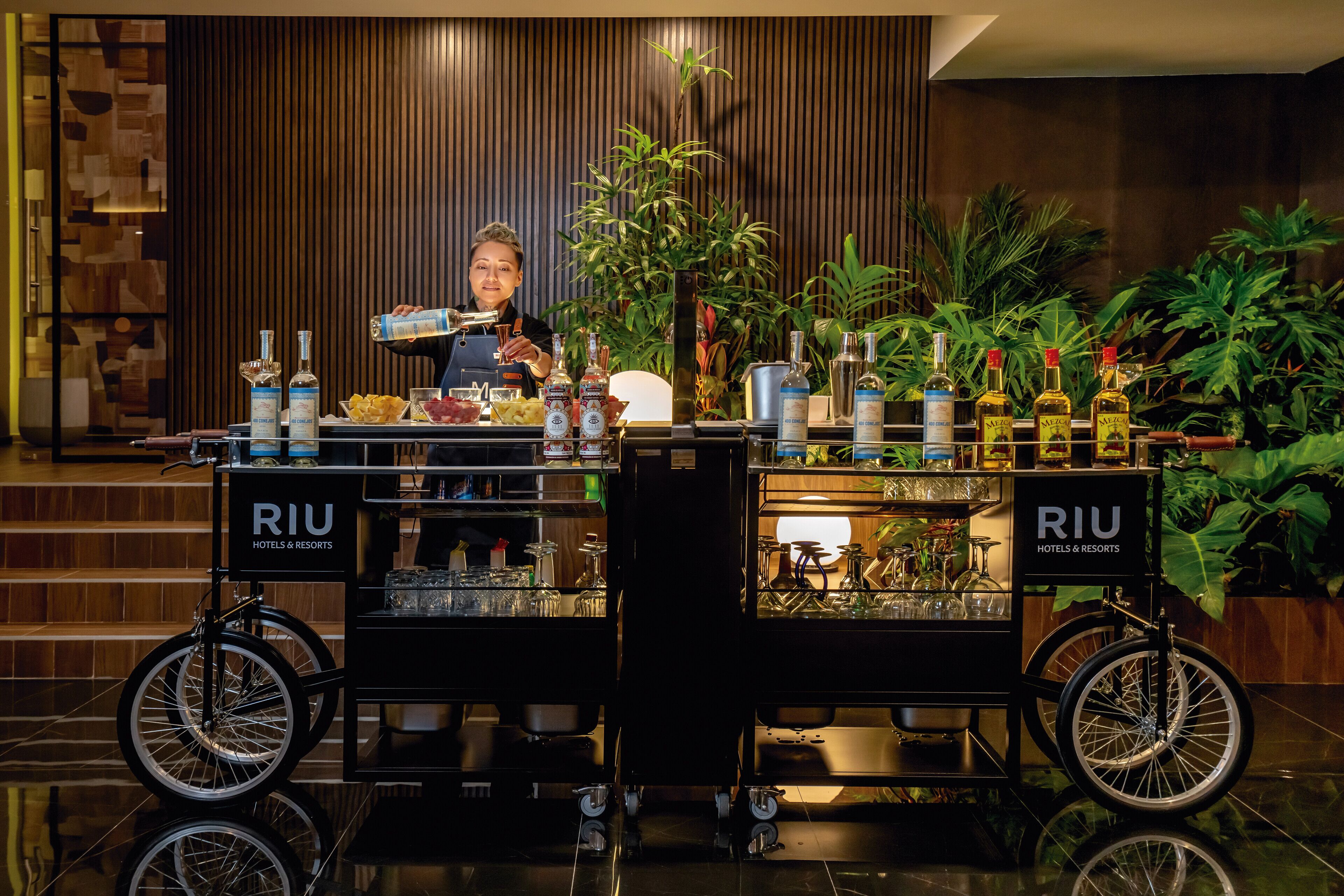 Un espace bar avec un barman, un chariot rempli de bouteilles et une enseigne indiquant « RIU HOTELS & RESORTS ».