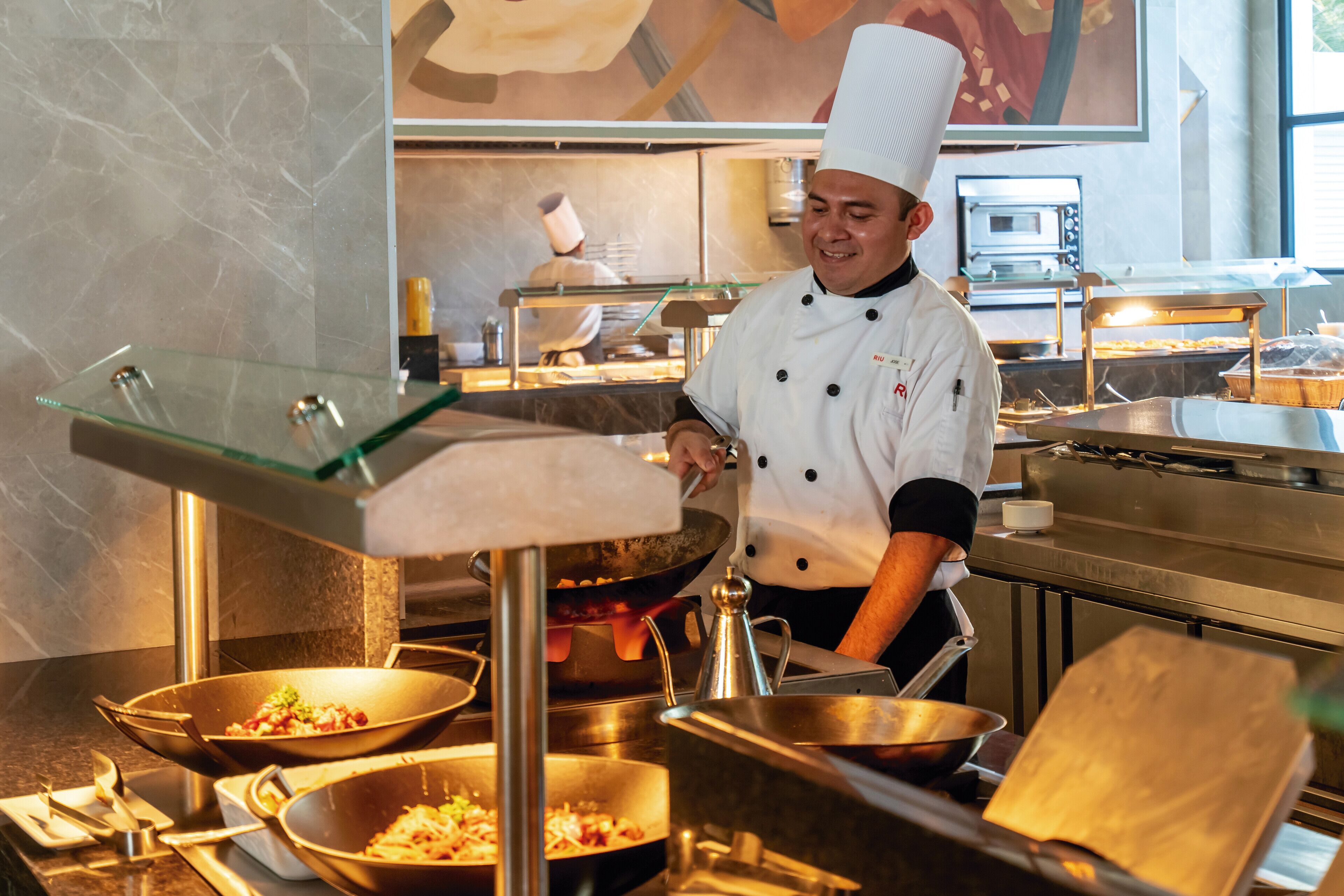 Un chef en una cocina profesional con electrodomésticos de acero inoxidable y una vitrina de vidrio.
