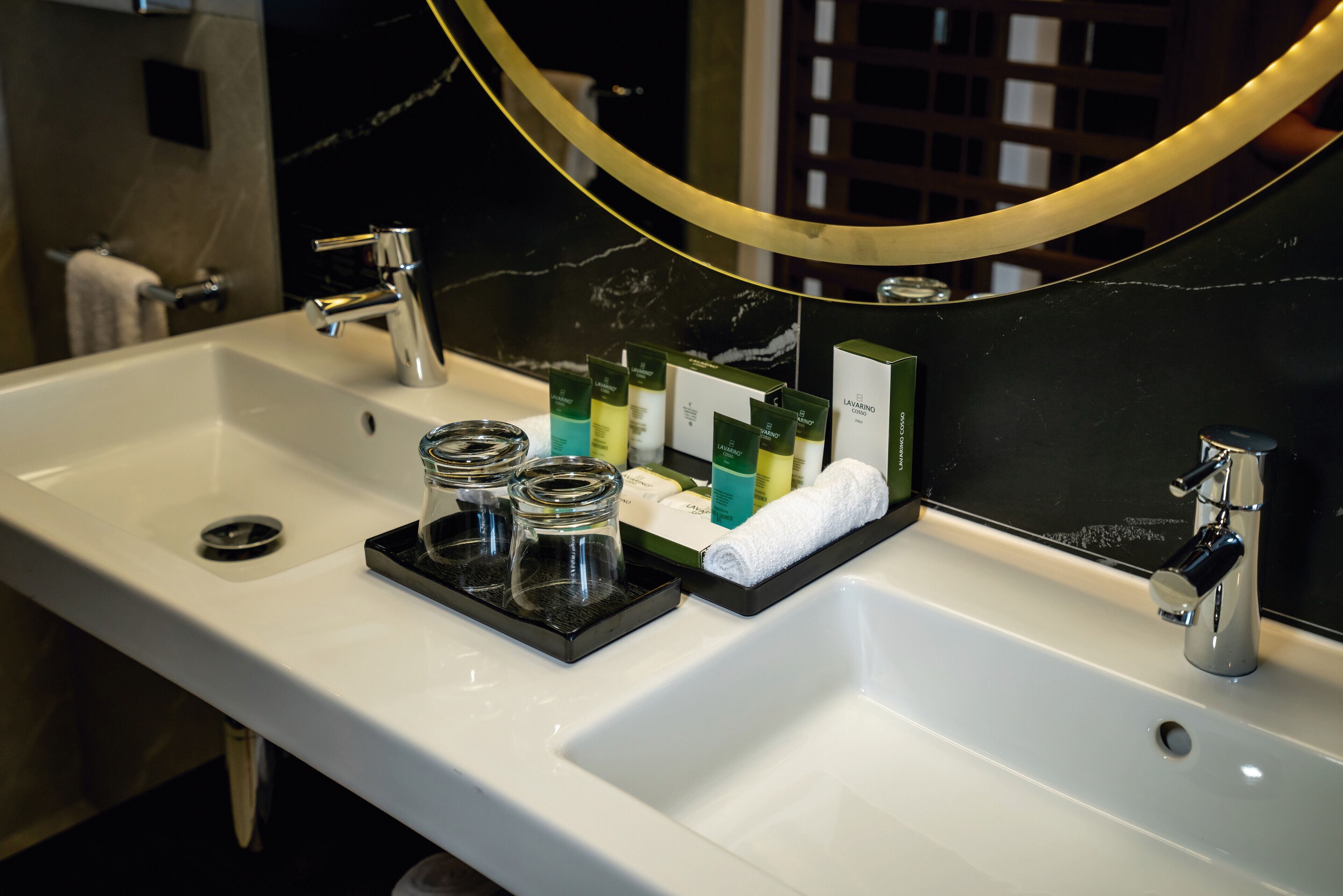 A hotel bathroom with a white sink, two faucets, and a mirror with a gold frame.