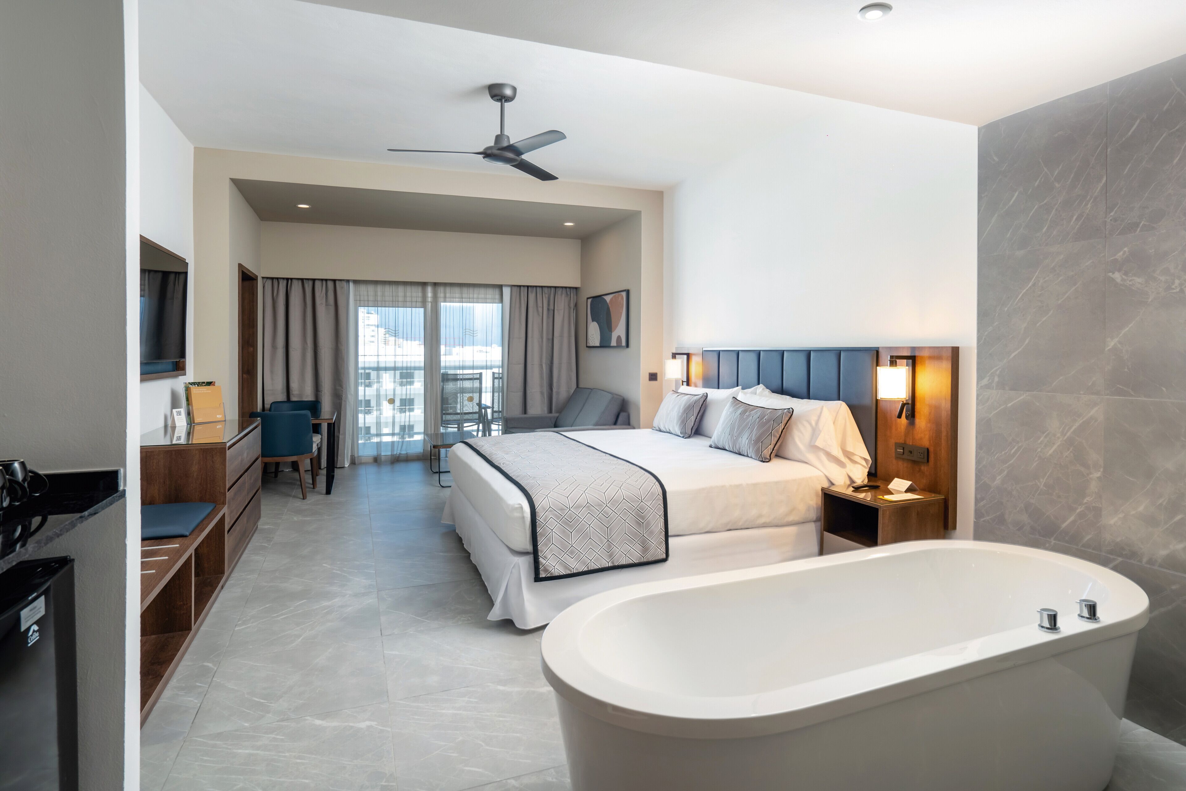 A modern hotel room with a large bed, a separate sitting area, a bathtub, and a desk with a chair.