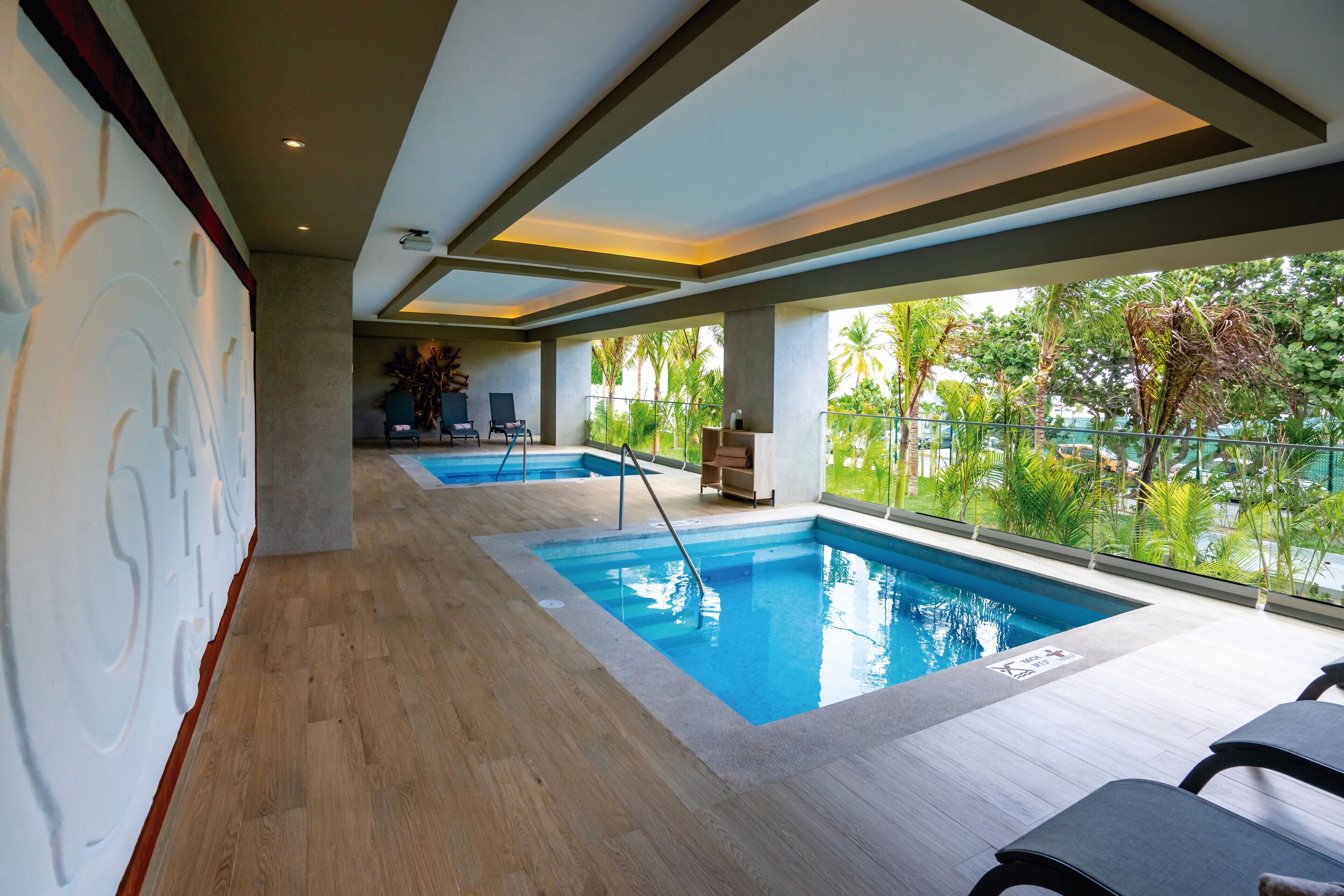 A modern indoor swimming pool area with two pools, wooden flooring, and large windows offering views of greenery.