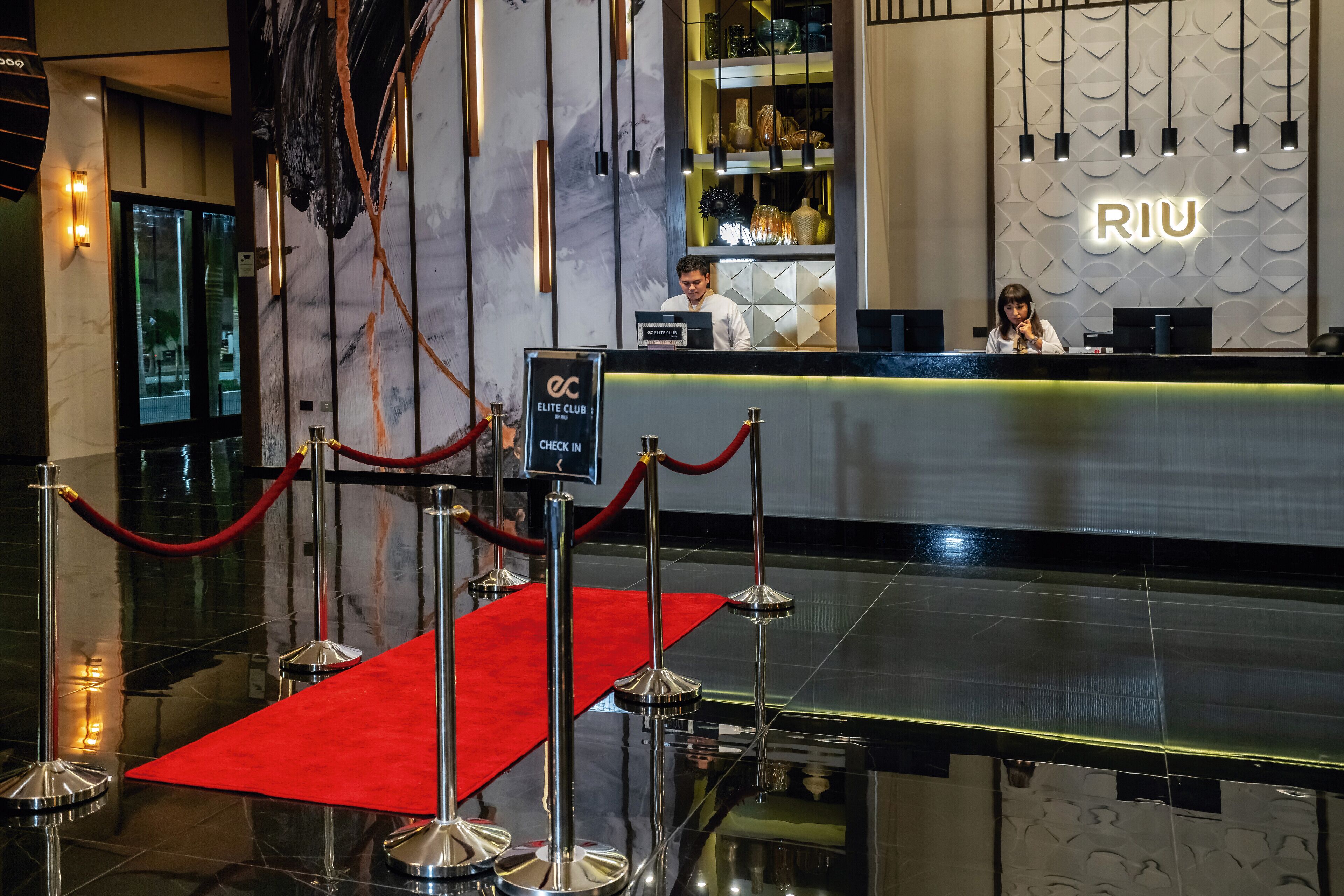 Un'area reception di un hotel con tappeto rosso, banco di registrazione e una parete con il nome 'RIU'.