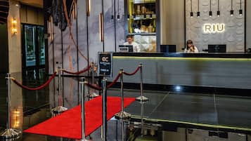 Un'area reception di un hotel con tappeto rosso, banco di registrazione e una parete con il nome 'RIU'.