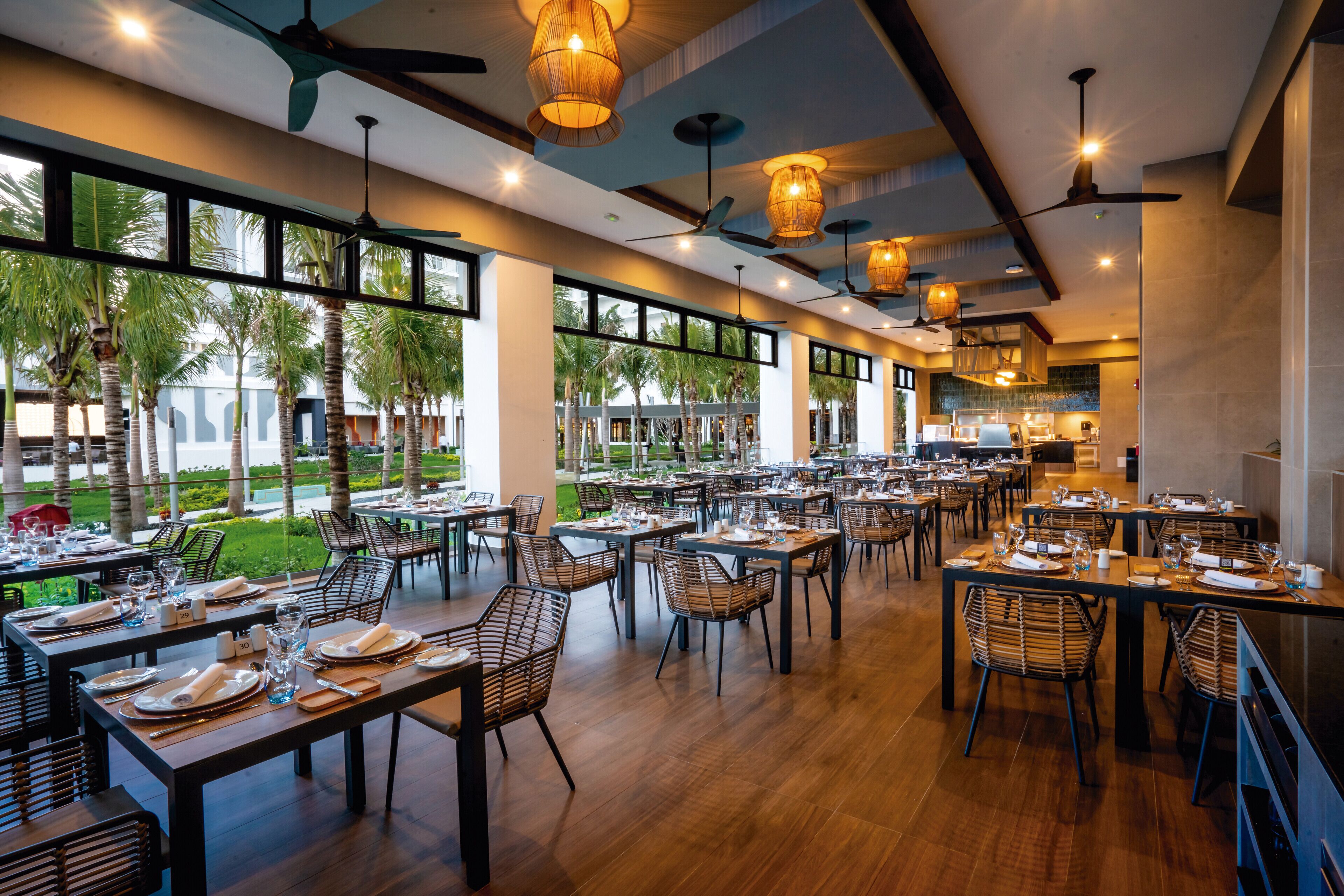 Un restaurant spacieux avec de hauts plafonds, de grandes fenêtres et une vue sur des palmiers. Les tables sont dressées avec soin, avec assiettes, verres et couverts.