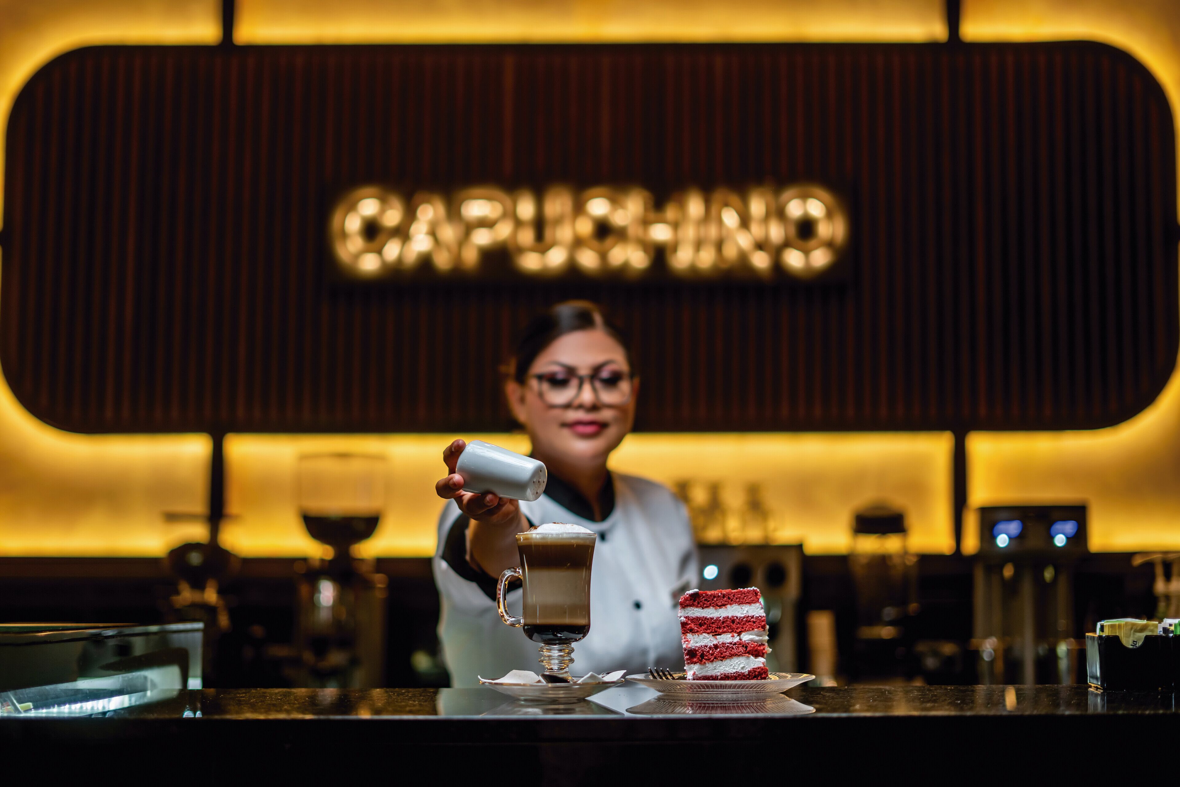 A barista at a coffee bar with a sign reading "CAPPUCCINO" in the background.