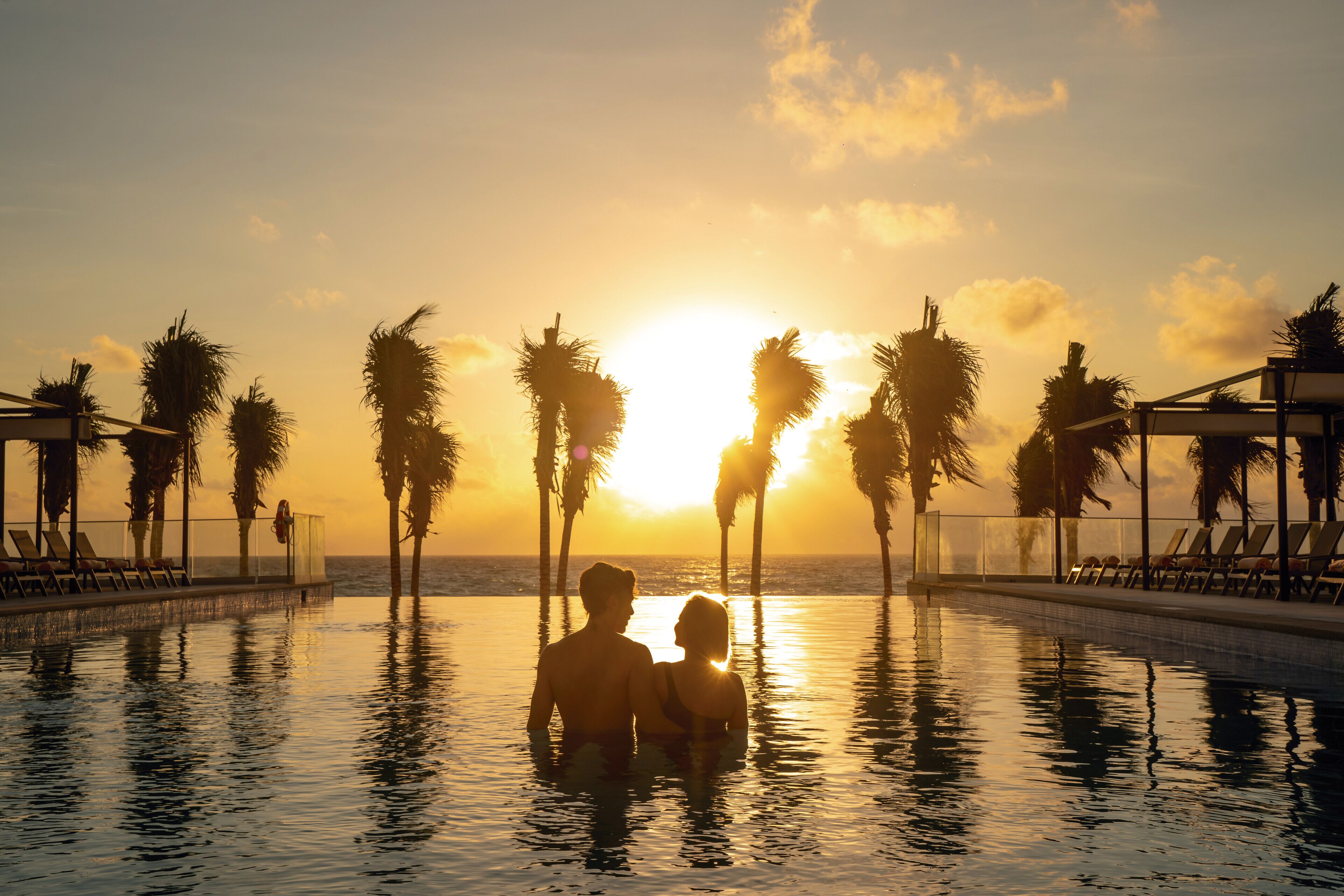 Una pareja en una piscina al atardecer, con palmeras y sillones de descanso alrededor.