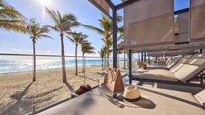 A beachside room with a view of the ocean, palm trees, and a sun lounger area.