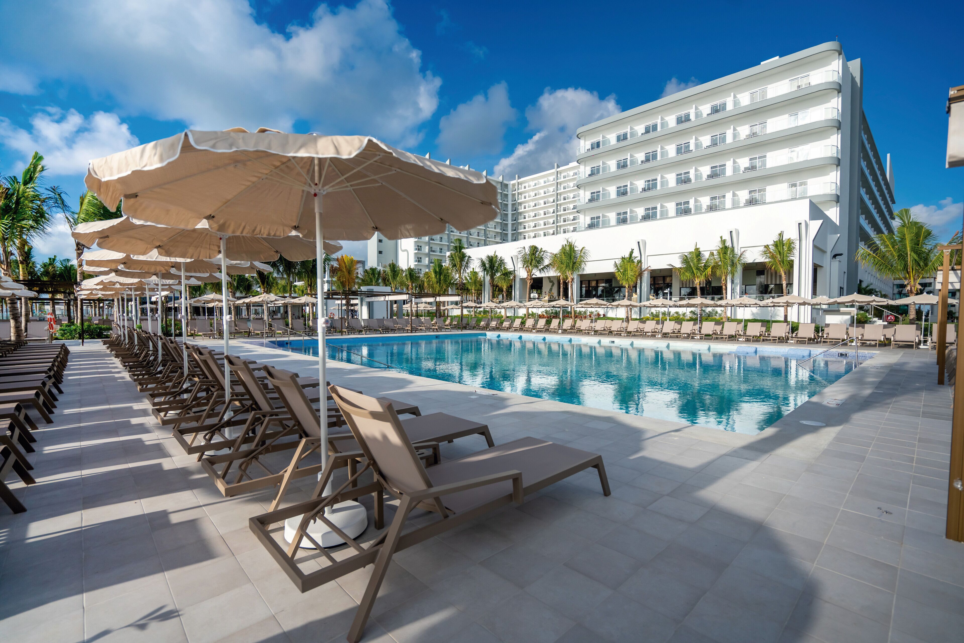 A poolside area with sun loungers and umbrellas, a large pool, and a white hotel building in the background.