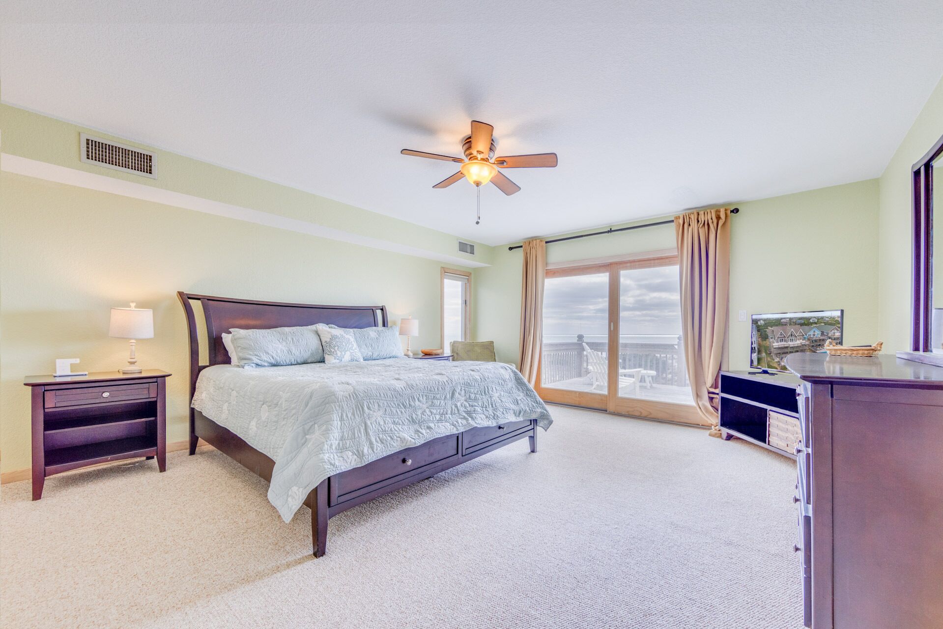 Une chambre à coucher comprenant un lit, des tables de chevet, un ventilateur de plafond, une grande fenêtre avec des rideaux et une vue sur l’extérieur.