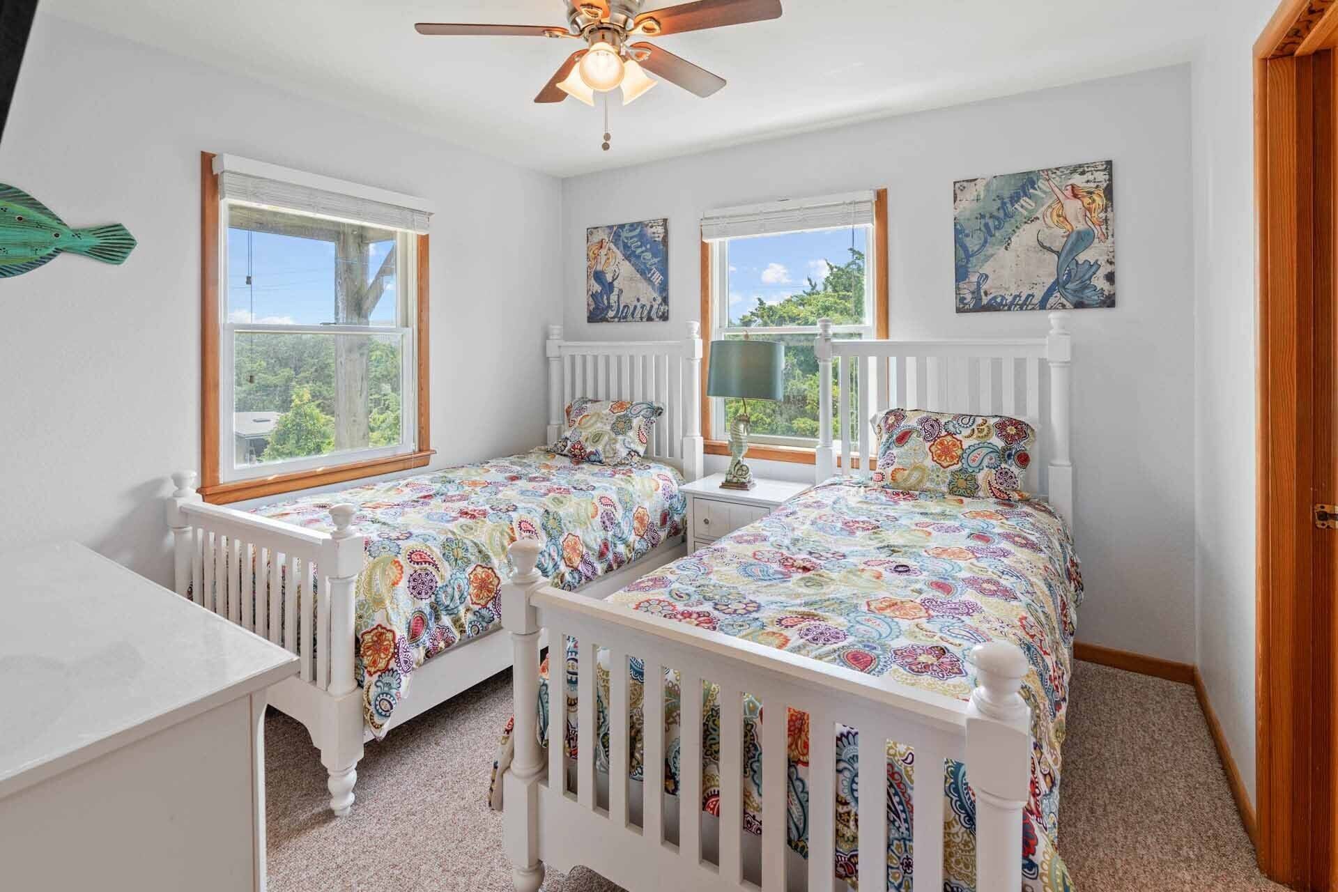 A bedroom with two single beds, a white dresser, a ceiling fan, and a window with a view of greenery.