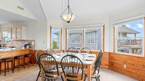 Espaço de refeições com mesa e cadeiras de madeira, janelas amplas e vista para uma casa.