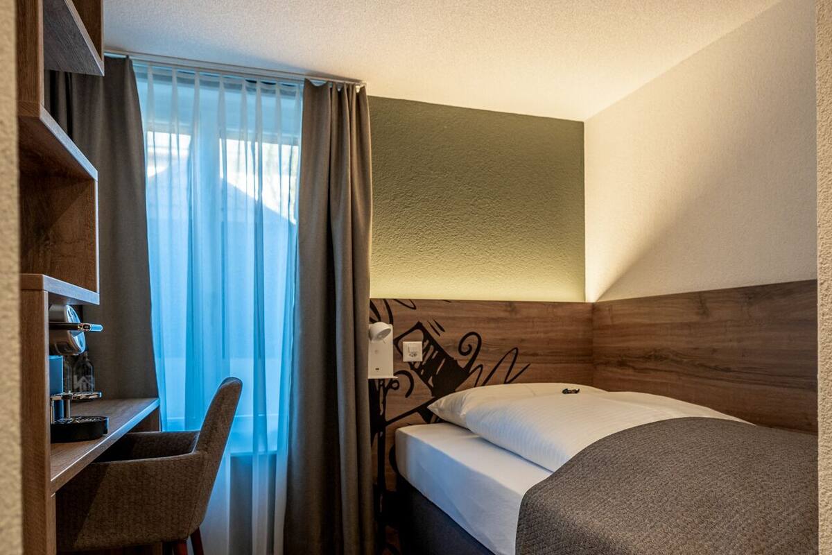 A compact hotel room with a bed, a desk, a chair, and a window with curtains.