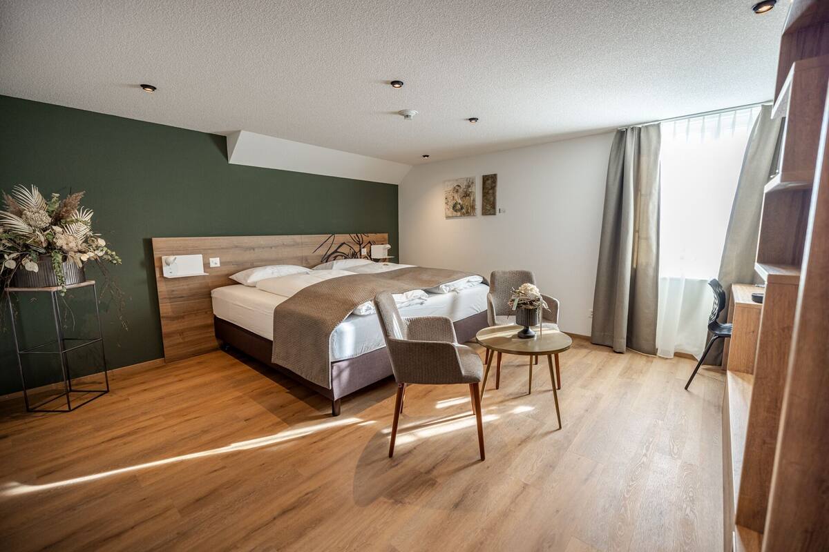 A hotel room with a bed, two chairs, a small table, a plant, and a window with curtains.