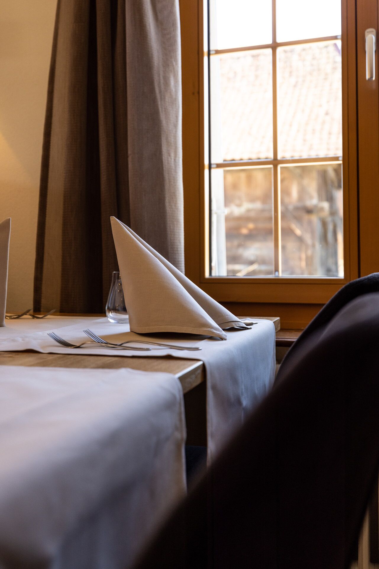 A dining table with a laptop, a glass, and cutlery, set against a window with curtains.