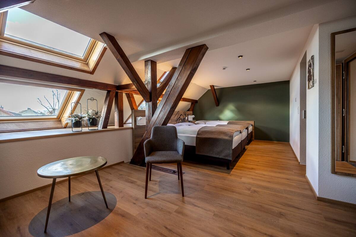 A modern bedroom with a skylight, wooden beams, a bed, a small table, and two chairs.