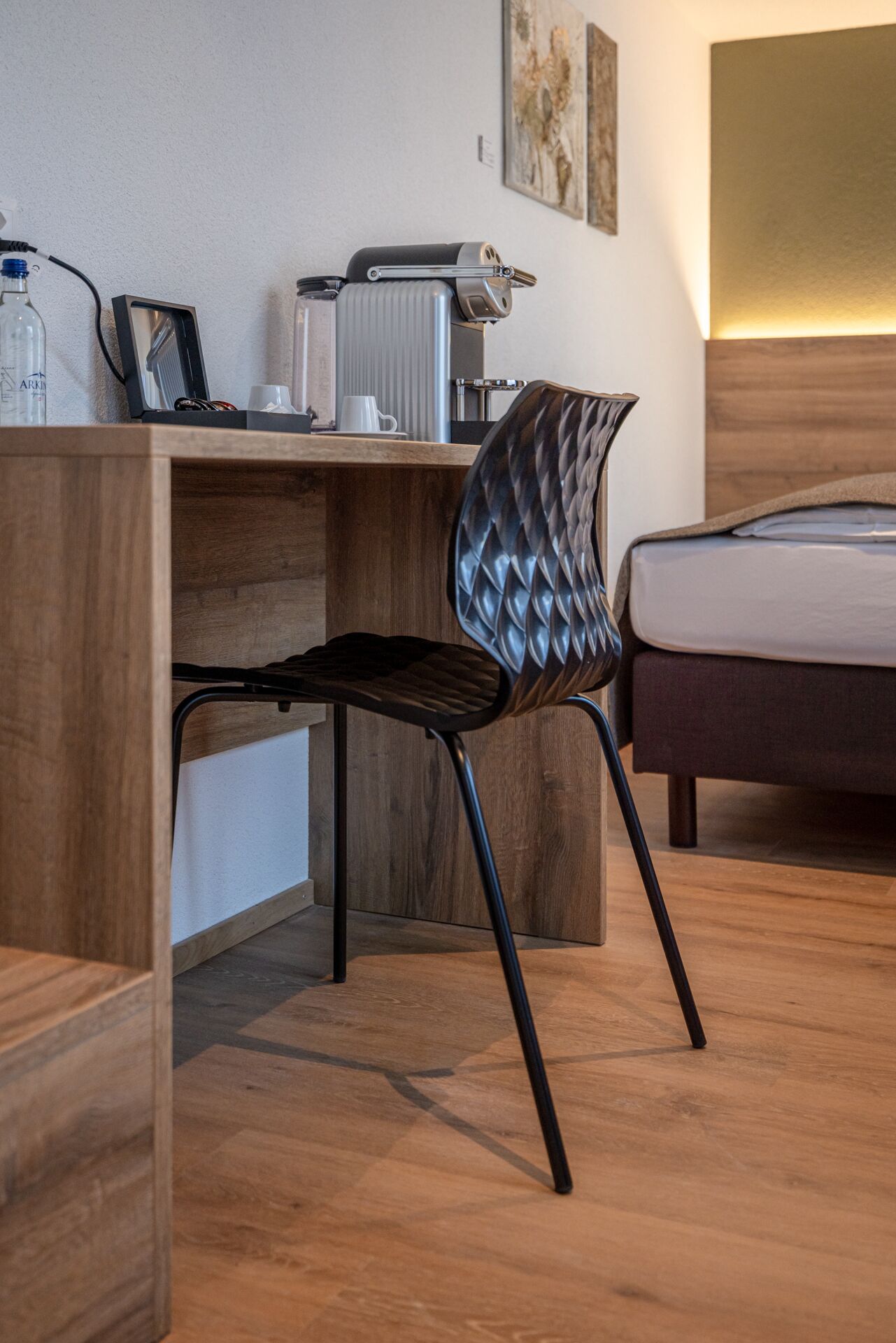 A modern hotel room with a wooden desk, a black chair with a unique design, a bed with a wooden headboard, and a radiator on the wall.