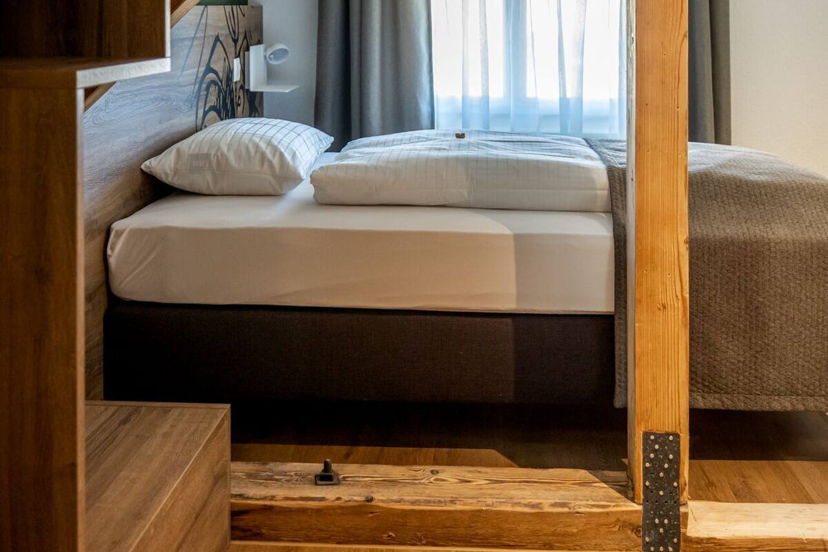 A bedroom with a wooden staircase leading to a loft area, a bed with white bedding, and a window with sheer curtains.