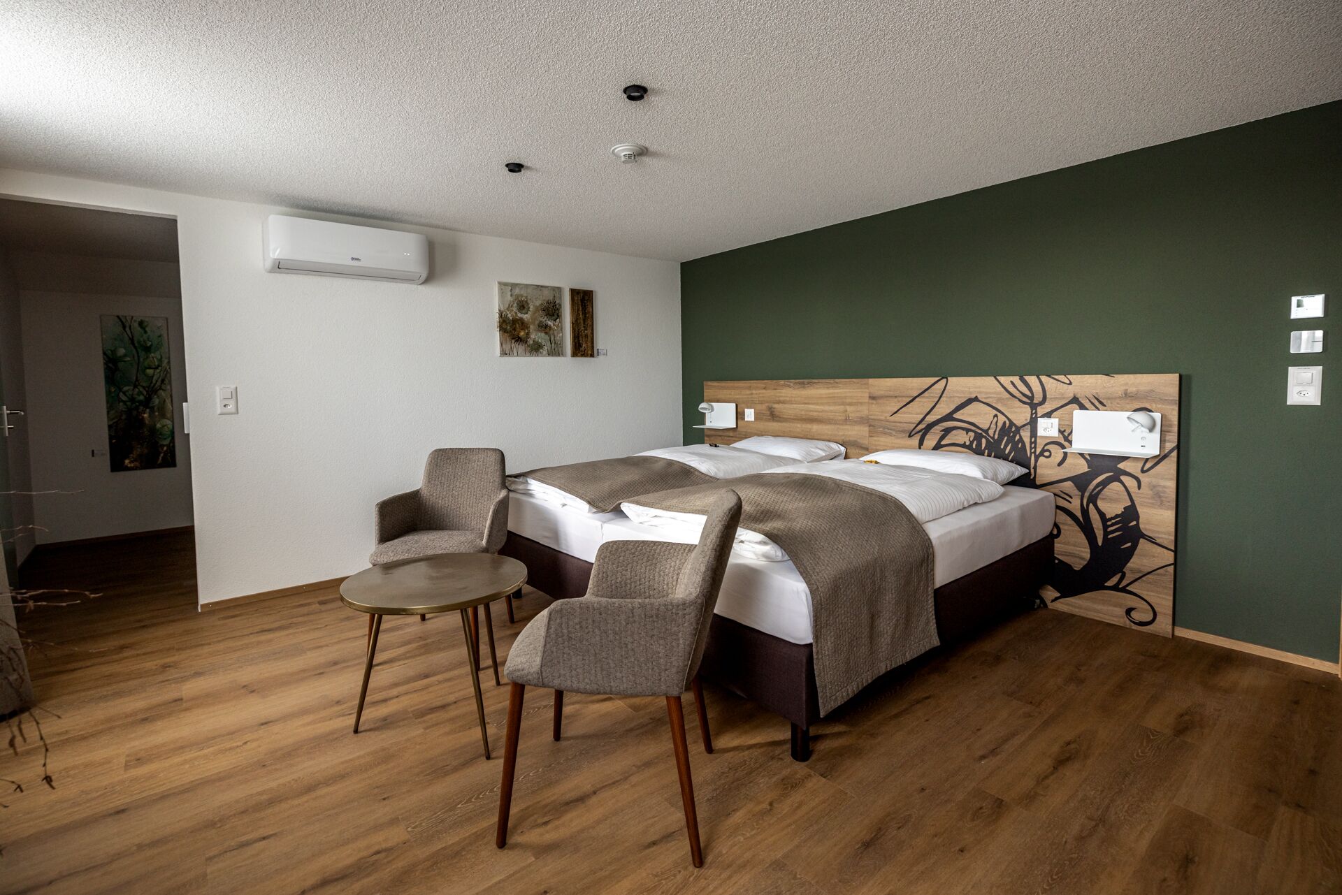 A bedroom with a bed, two chairs, a small table, an air conditioner, and a wall-mounted light fixture.