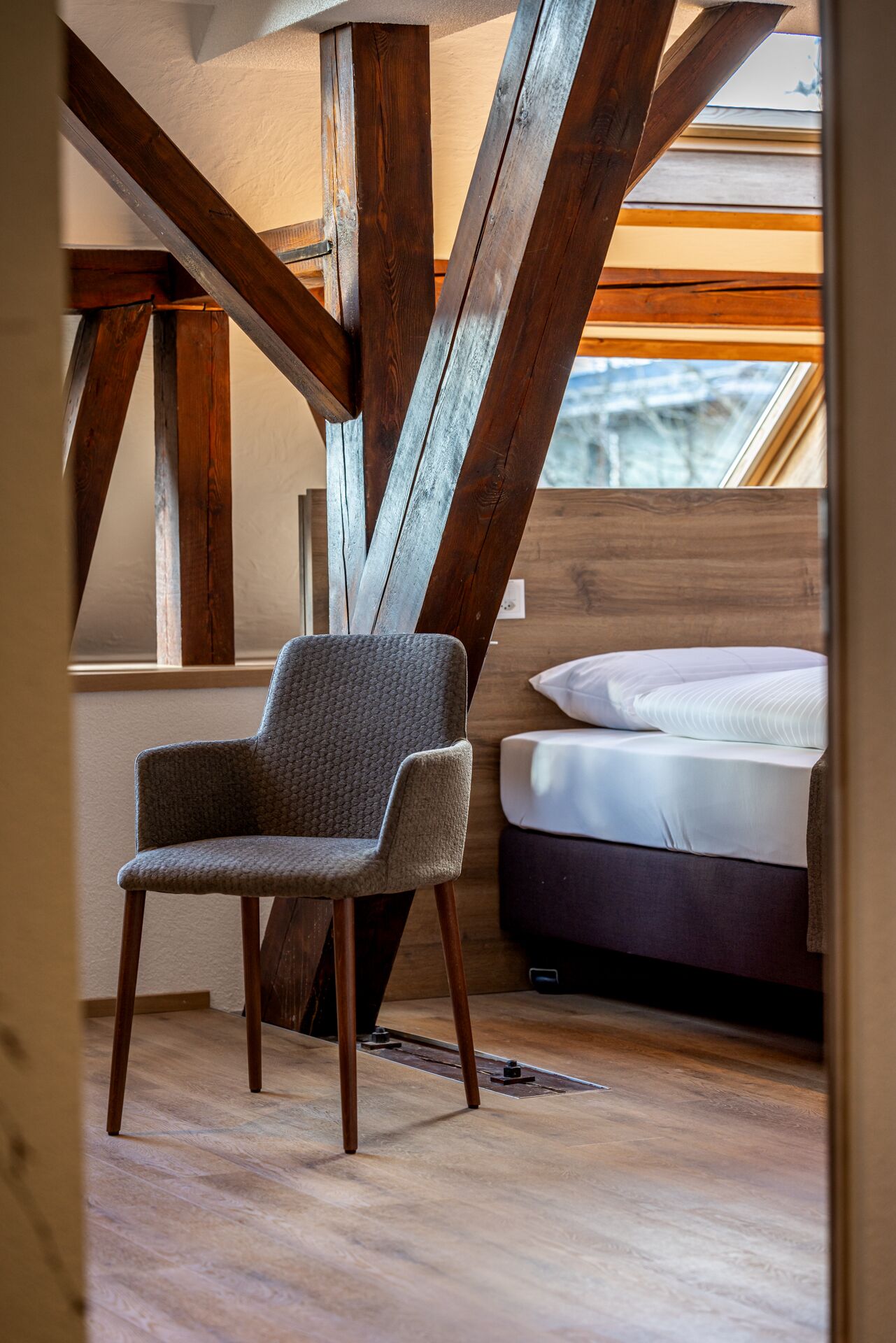 Une pièce avec un plafond haut, des poutres apparentes en bois, un fauteuil recouvert d’un tissu gris, un lit avec une literie blanche et une vue par la fenêtre.