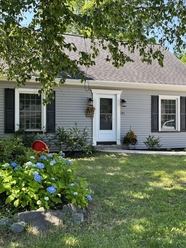 A single-story house with a gray siding exterior, a white front door, and black shutters. There is a number '911' on the house, a small porch with a red chair, and a garden with blue hydrangea flowers and green foliage.
