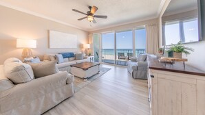 Un salon spacieux doté d’un ventilateur de plafond, d’une grande fenêtre offrant une vue sur l’océan et d’un balcon aménagé avec du mobilier d’extérieur.