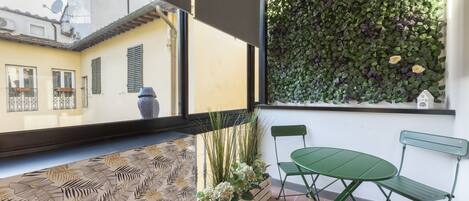 Un balcón con suelo estampado, una mesa y silla verde, una maceta de madera con plantas, y una pared cubierta de follaje.