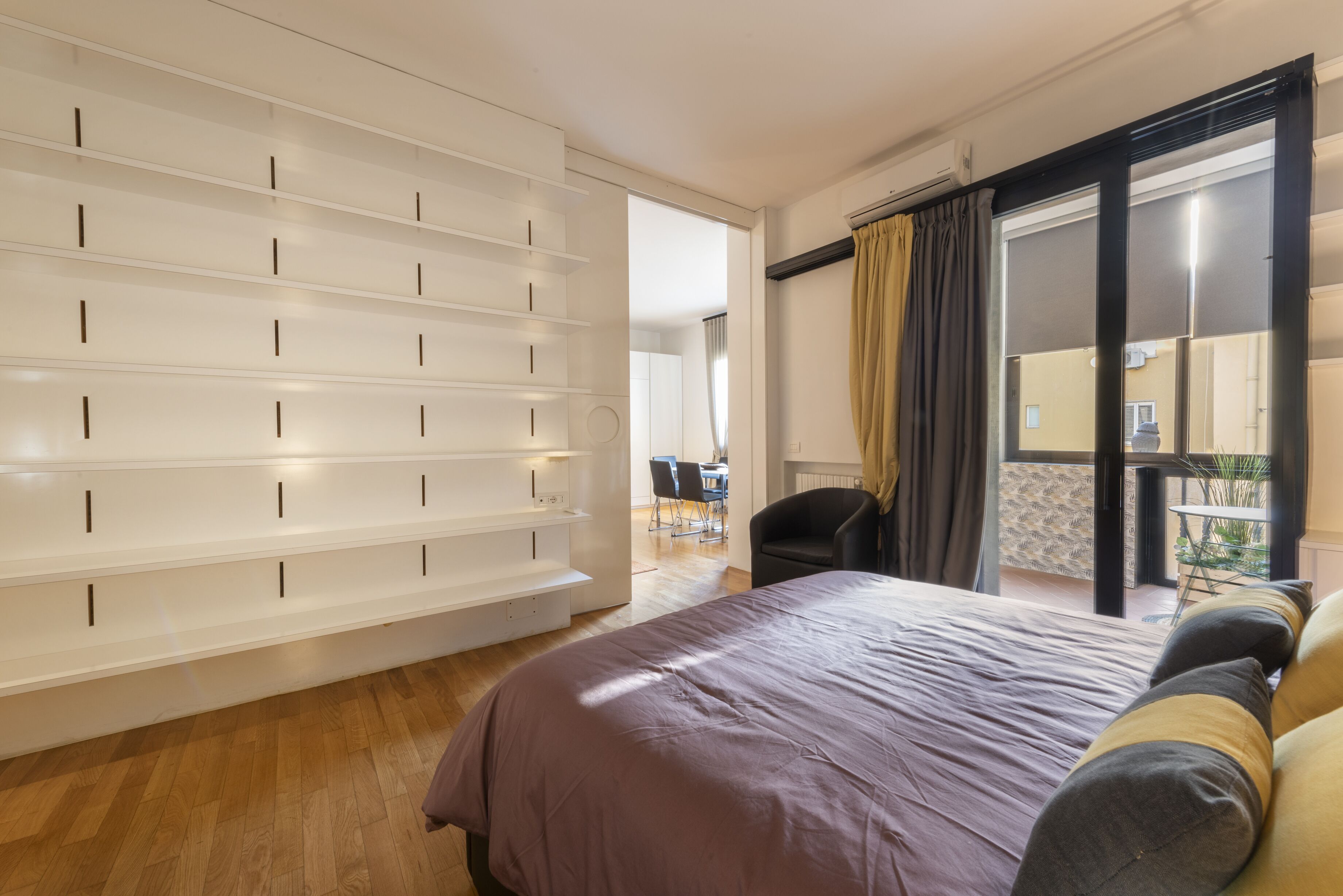 Une chambre avec un grand lit, une armoire avec de nombreuses étagères, une chaise et un balcon avec vue.