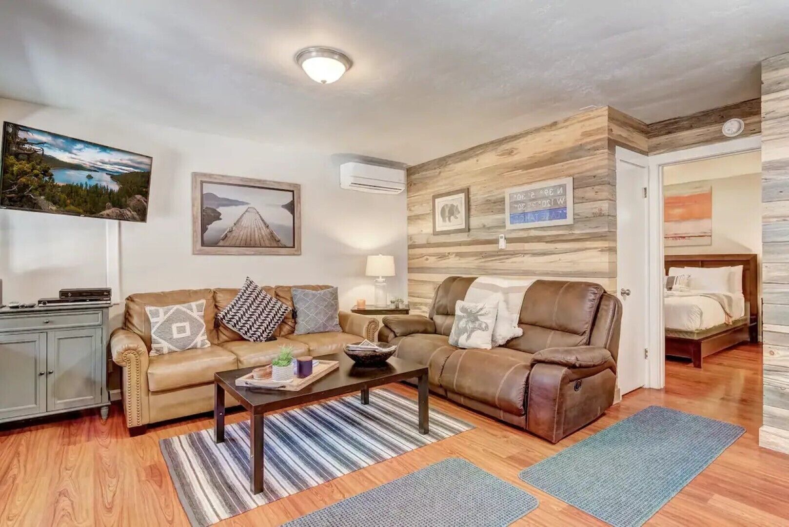 A living room with a sofa, a coffee table, and a TV. There is a wooden wall with pictures and a door leading to a bedroom.