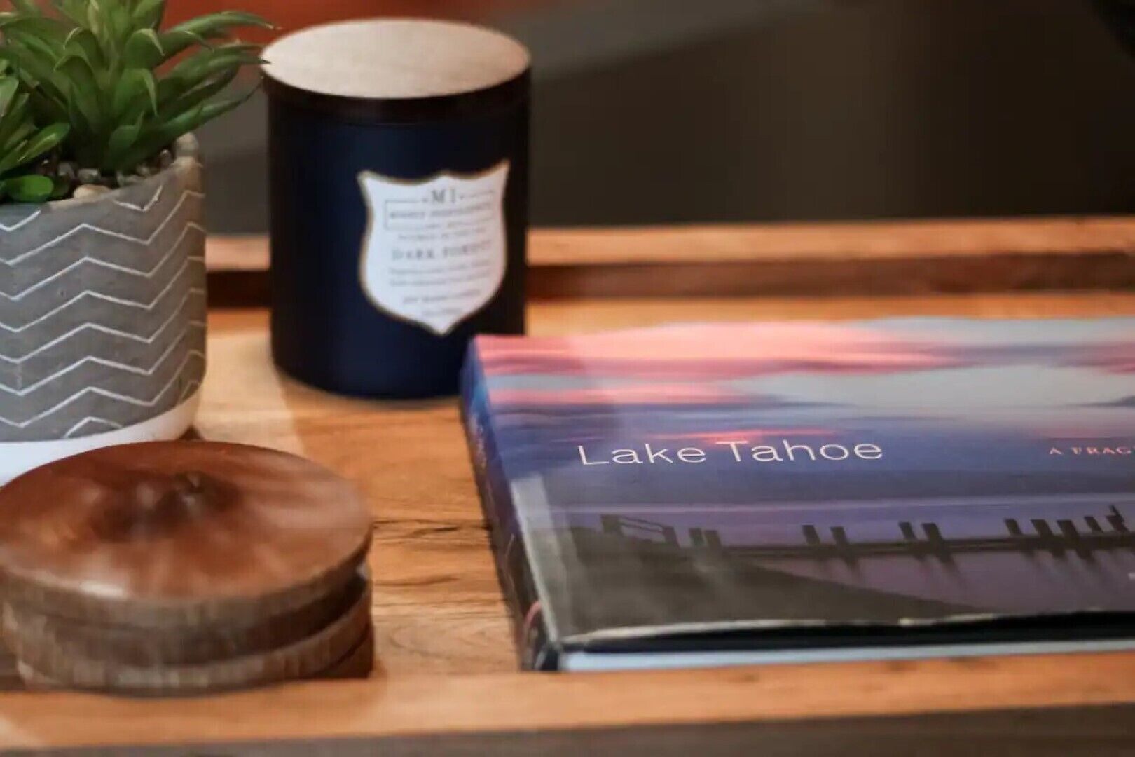 Een boek met de titel 'Lake Tahoe' op een houten ondergrond, een donkere kaars en een kleine plant in een pot.