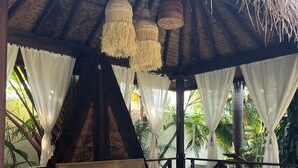 Un lit en bois sous un toit de chaume, avec des lampes suspendues et des rideaux blancs.