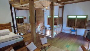 Une pièce spacieuse avec un plafond en bois, un lit avec une literie blanche, une table à manger avec des chaises, et une vue sur un balcon doté de balustrades en bois.