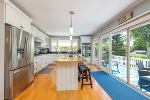 A modern kitchen with stainless steel appliances, a central island with stools, and large windows overlooking a deck. - Sand Dollar - Dog-friendly, Steps to the Ocean, Stunning Sunsets (Harpswell)