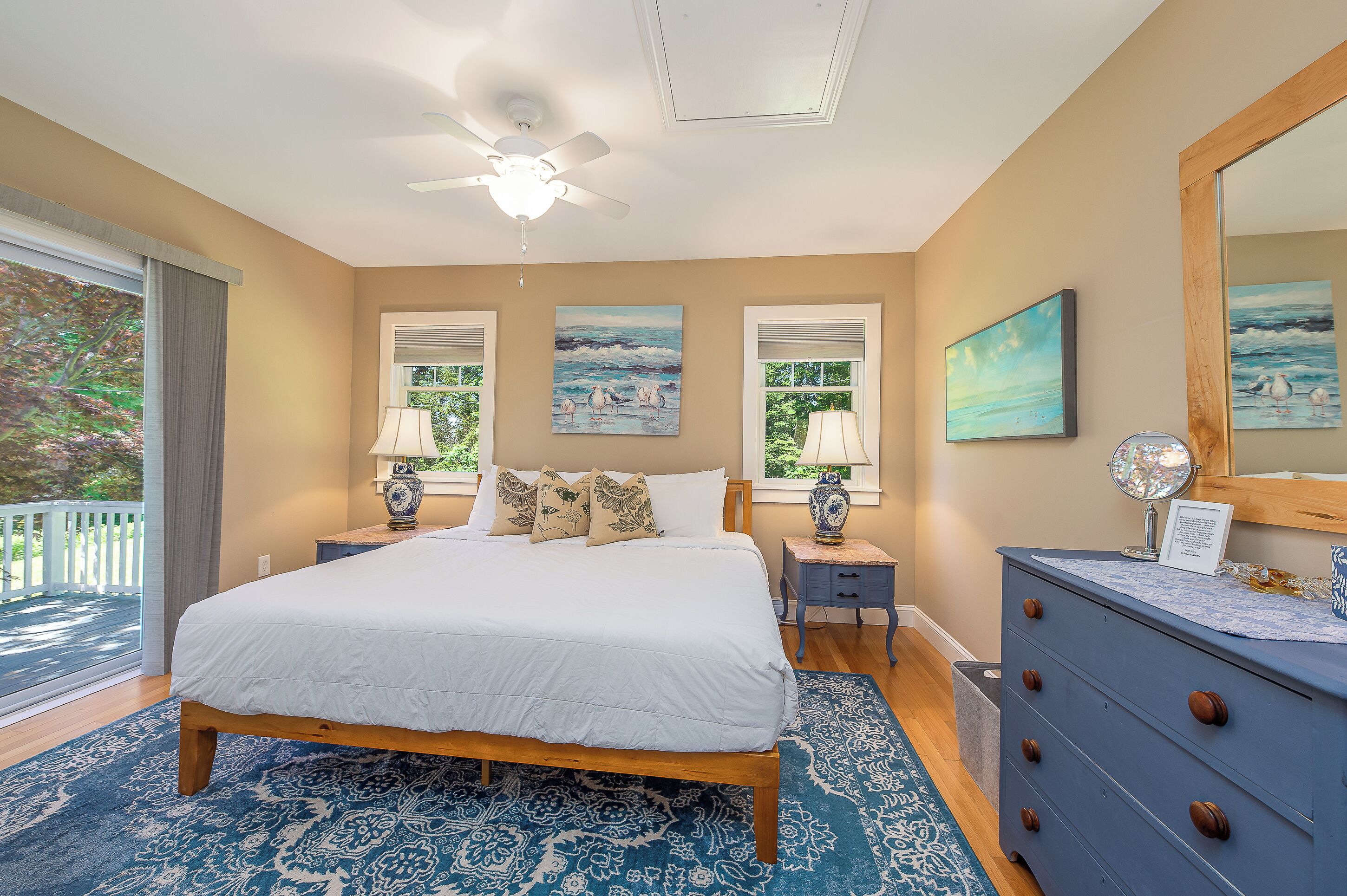 A bedroom with a large bed, two bedside tables, a dresser, a ceiling fan, and a view of the outdoors through windows.