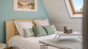 Une chambre à coucher avec un lit, des oreillers, une table de chevet, un tableau encadré au mur et une lucarne.