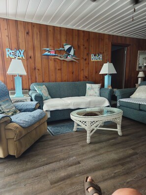 A room with wooden panel walls, a blue sofa, a beige armchair, a glass coffee table, and two lamps.