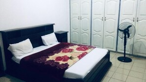 Ein Schlafzimmer mit einem Bett, weißen Kissen, einer blumigen Tagesdecke, einem dunklen Kopfteil, einem Nachttisch, einem Standventilator und einem Kleiderschrank mit mehreren Türen.