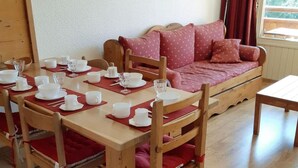 A dining area with a wooden table set for four, surrounded by wooden chairs with red cushions. A wooden sofa with red cushions is against the wall, and a floor lamp is visible. - 100m from ski slopes, 5th floor, view mountain, balcony, parking, tv, ski locker, 40m², Les 2 Alpes (Les Deux Alpes)