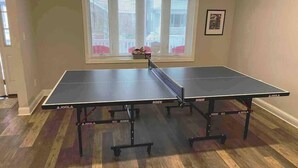 A ping pong table in a room with wooden flooring, a window with blinds, and a framed picture on the wall. - All new beach block Bartram Beach Tower - 8bd home (Atlantic City)