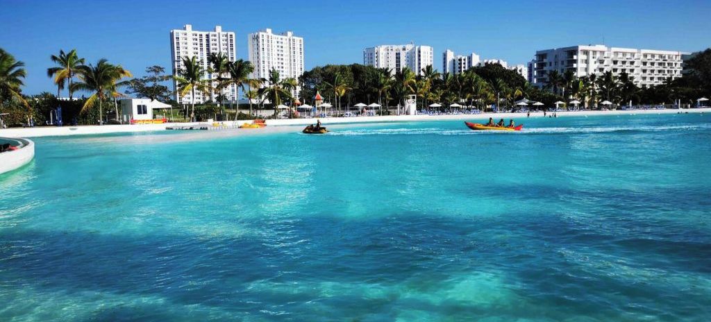A beach with clear turquoise water, palm trees, and high-rise buildings in the background.