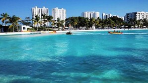 Une plage avec une eau turquoise et limpide, des palmiers et, en arrière-plan, des immeubles de grande hauteur.