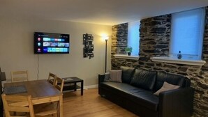 A living room with a stone wall, a black leather sofa, a wooden dining table with chairs, and a flat-screen TV mounted on the wall. - Cozy 1-bedroom in Bauer Terrace next to Citadel (Halifax)
