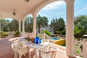 Un área de comedor al aire libre techada con sillas blancas y una mesa dispuesta para dos, con vista a una piscina y follaje.