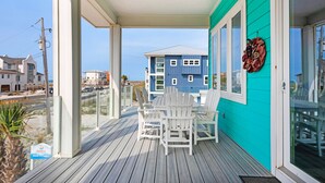 Een overdekt buitengebied met een houten terras, witte Adirondack-stoelen en een uitzicht op een blauw gebouw op de achtergrond.